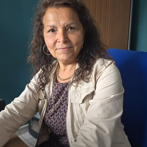 souriante assise à son bureau dans un environnement de travail, avec des dossiers et cartons à l’arrière-plan.